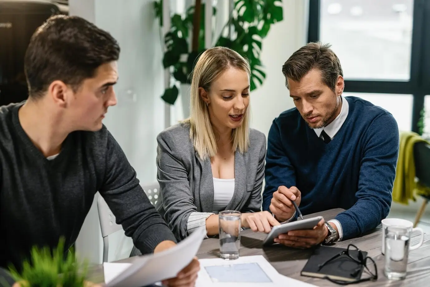 A real estate agent and a young couple are reviewing housing plans on a touchpad during a home meeting.