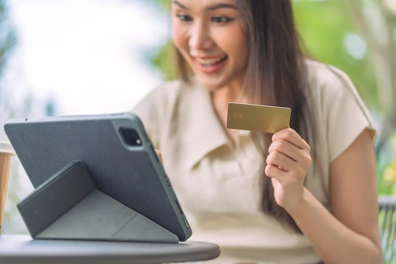 An Asian woman sits at an outdoor café on a weekend vacation, shopping online on her smartphone and making a mobile payment with a credit card.
